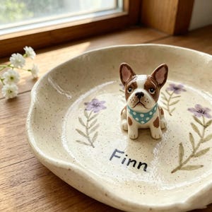 May include: A ceramic dog-themed dish with a French Bulldog figurine. The off-white dish features hand-painted purple flowers and green leaves. The dog figurine has brown and white markings and a blue polka-dot bandana. The name "Finn" is written on the dish.