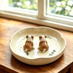 May include: A ceramic pet bowl with a cream-colored base, featuring two brown and white bulldog figurines. The bowl has a scalloped edge and is decorated with small white daisy and green leaf designs. The bowl is sitting on a wooden surface.