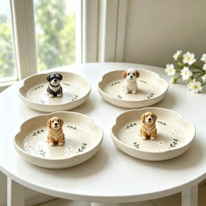 May include: Four beige ceramic dishes with scalloped edges, each featuring a different dog figurine. The dishes are decorated with a floral pattern of white daisies and green leaves. The dogs include a black and white Schnauzer, a white and brown Shih Tzu, and two Golden Retrievers.