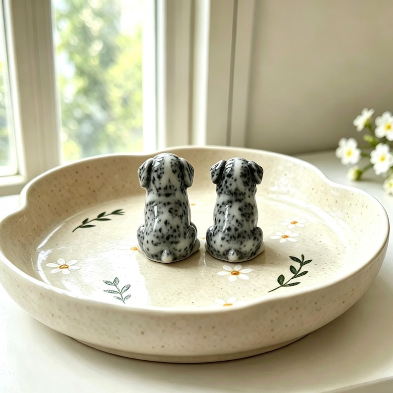 May include: Two ceramic Dalmatian dog figurines with black spots sit on a beige, scalloped-edge plate decorated with white daisies and green leaves. The plate has a speckled texture and is on a white surface.