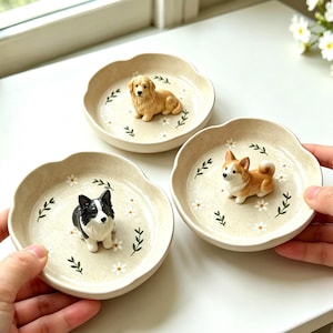 May include: Three ceramic dog-themed dishes. Each dish is beige with a scalloped edge and decorated with white flowers and green leaves. Each dish features a different dog breed figurine: a golden retriever, a corgi, and a border collie.