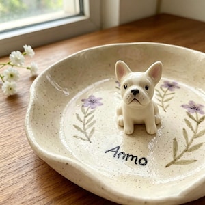 May include: A cream-colored ceramic dish with a white French bulldog figurine in the center. The dish is decorated with purple floral designs and the word "Ammo" in black script. The dish has a speckled texture.
