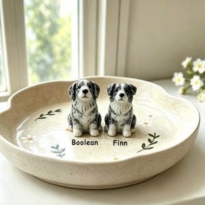 May include: Two ceramic dog figurines with black and white speckled patterns sit on a beige, scalloped-edge dish decorated with daisies and green leaves. The dish has the names "Boolean" and "Finn" printed on it.