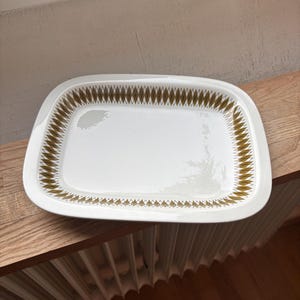 May include: A rectangular white serving platter with a gold diamond pattern around the rim. The platter is made of ceramic and has rounded corners. The design is symmetrical and the platter is sitting on a wooden surface.