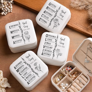 May include: White jewelry travel cases with inspirational text and floral designs. One case is open, revealing various rings, necklaces, and earrings. The cases are arranged on a brown surface, with decorative elements.