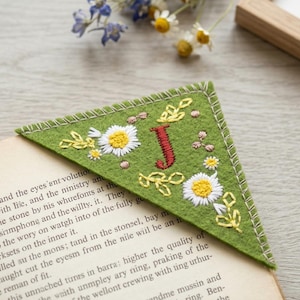 Puede incluir: Marcapáginas triangular de fieltro verde con bordados de margaritas, hojas y la letra "J" roja. El marcapáginas, con un borde cosido, está colocado en la esquina de un libro.