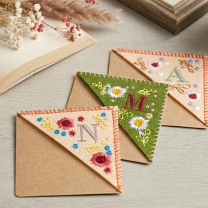 Könnte beinhalten: Drei handgefertigte Eck-Lesezeichen aus Filz mit gestickten Blumenmustern und Initialen. Eines ist grün mit einer weißen Gänseblümchen und dem Buchstaben "M", ein anderes ist beige mit dem Buchstaben "A" und das dritte ist beige mit dem Buchstaben "N".