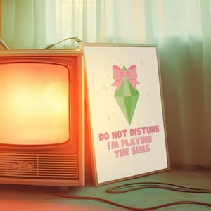 May include: A white poster with a green diamond shape with a pink bow on top. The text on the poster reads "Do Not Disturb I'm Playing The Sims".
