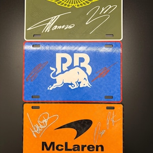 May include: Three decorative license plates featuring racing team logos and signatures. The top plate is olive green with an Aston Martin logo and a signature. The middle plate is blue with a white bull logo and a signature. The bottom plate is orange with a McLaren logo and signatures.