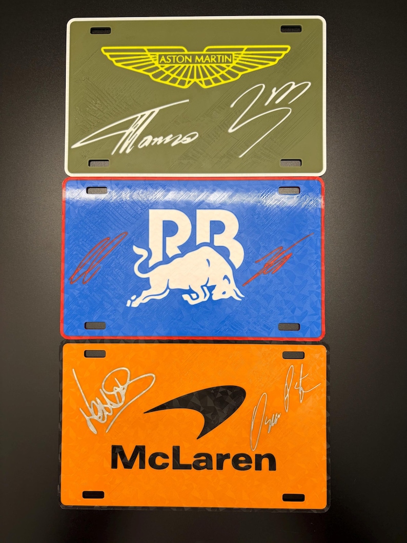 May include: Three decorative license plates featuring racing team logos and signatures. The top plate is olive green with an Aston Martin logo and a signature. The middle plate is blue with a white bull logo and a signature. The bottom plate is orange with a McLaren logo and signatures.