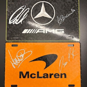 May include: Two license plates, one black with a Mercedes-AMG logo and signatures, the other orange with a McLaren logo and signatures. Both plates have a textured pattern.