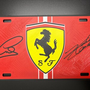 May include: Red license plate featuring the Ferrari logo, a black prancing horse on a yellow shield, with the letters "SF" below. The plate has two signatures and a textured background.