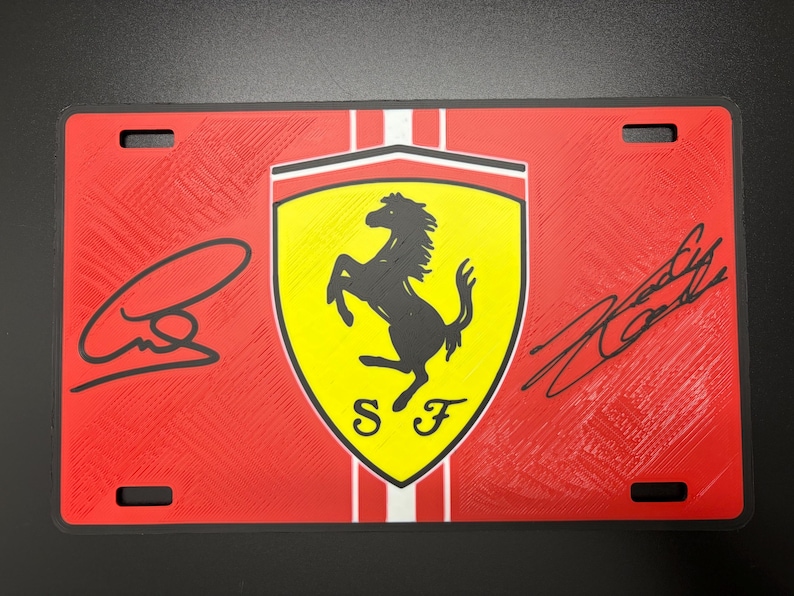 May include: Red license plate featuring the Ferrari logo, a black prancing horse on a yellow shield, with the letters "SF" below. The plate has two signatures and a textured background.