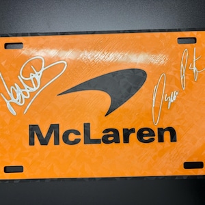 May include: Orange McLaren license plate with black accents and two signatures. The McLaren logo is in the center, with the word "McLaren" below it. The plate has four mounting holes.