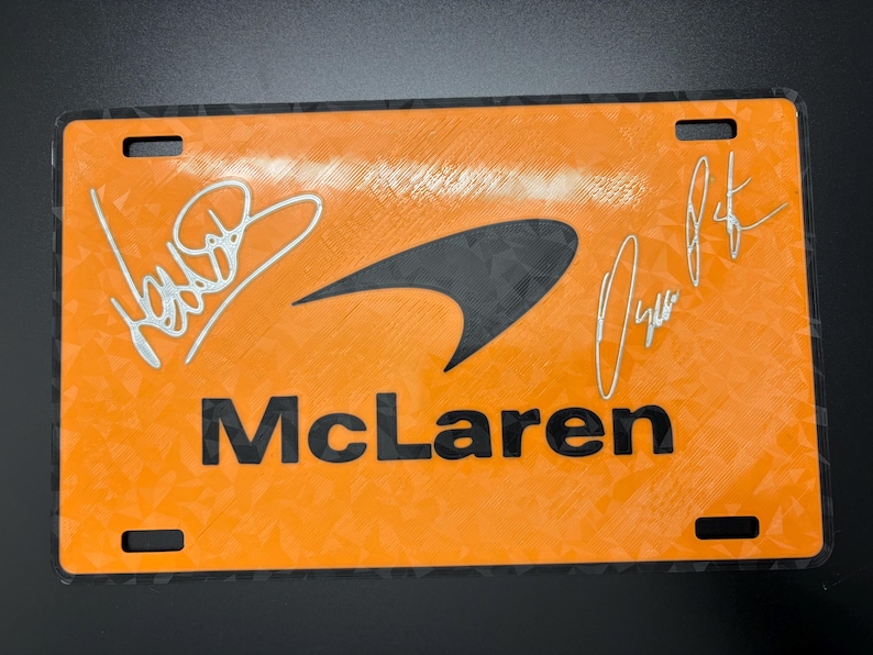May include: Orange McLaren license plate with black accents and two signatures. The McLaren logo is in the center, with the word "McLaren" below it. The plate has four mounting holes.