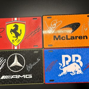 May include: Four license plates featuring racing team logos and signatures. The plates display the Ferrari logo on red, McLaren on orange, AMG on black, and Red Bull on blue. Each plate has a unique design and color scheme.