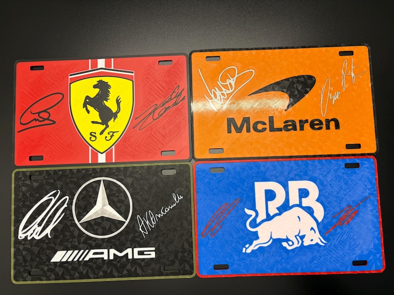 May include: Four license plates featuring racing team logos and signatures. The plates display the Ferrari logo on red, McLaren on orange, AMG on black, and Red Bull on blue. Each plate has a unique design and color scheme.