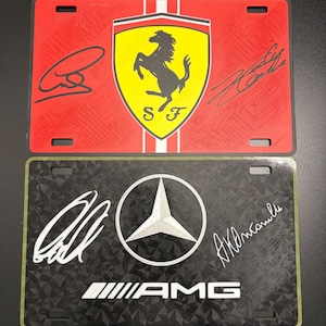 May include: Two license plates. The top plate is red with the Ferrari logo and signatures. The bottom plate is black with the Mercedes-AMG logo and signatures. Both plates have mounting holes.