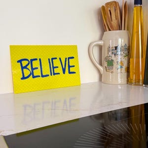 Peut inclure: Un panneau rectangulaire jaune à la surface texturée portant le mot BELIEVE écrit en lettres capitales bleu vif. Le panneau est placé à côté d'une chope à bière en céramique contenant des ustensiles de cuisine en bois et une bouteille d'huile.