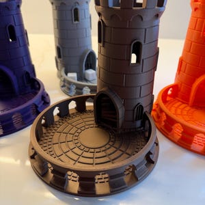 Könnte beinhalten: Eine Sammlung von 3D-gedruckten Miniatur-Burgtürmen in verschiedenen Farben: Lila, Grau, Braun und Orange. Jeder Turm hat ein zylindrisches Design mit gewölbten Fenstern und einer kreisförmigen Basis mit dekorativen Details. Die Türme sind wahrscheinlich für Tabletop-Spiele oder zur Präsentation.