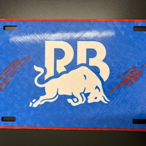 May include: Blue license plate with a white bull logo and the letters "RB". The plate has a red border and four mounting holes. Red streaks are on the left and right sides of the plate.