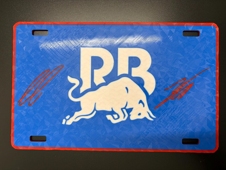 May include: Blue license plate with a white bull logo and the letters "RB". The plate has a red border and four mounting holes. Red streaks are on the left and right sides of the plate.
