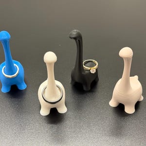 Può includere: Quattro portaanelli a forma di dinosauro in blu, bianco, nero e beige. Un anello d'oro con un diamante è sul supporto nero. I portaanelli sono su una superficie scura.