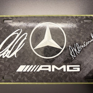 May include: Black license plate featuring the Mercedes-Benz logo, AMG text, and two signatures in white. The plate has a textured background and a green border. The Mercedes-Benz logo is a white star inside a white circle.