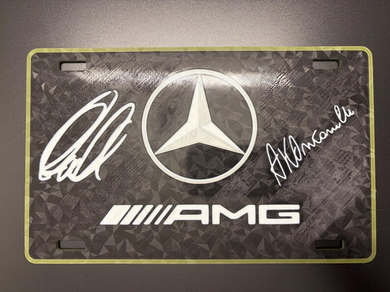 May include: Black license plate featuring the Mercedes-Benz logo, AMG text, and two signatures in white. The plate has a textured background and a green border. The Mercedes-Benz logo is a white star inside a white circle.