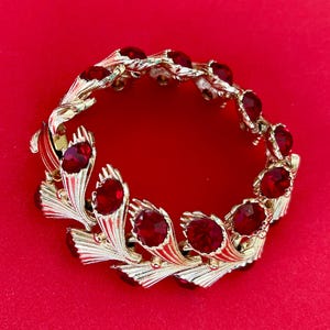 May include: A gold-toned bracelet with a leaf design, adorned with vibrant red gemstones. The bracelet is set against a red background, highlighting the contrast between the gold and the red stones. The bracelet is a piece of jewelry.