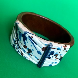 May include: A ceramic bangle bracelet with a white base and blue and brown splatter paint design. The bangle is open on one side and has a dark brown interior.