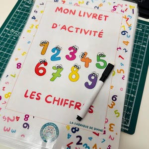 Peut inclure: Un livret d'activités coloré avec le titre "MON LIVRET D'ACTIVITÉ" et les mots "LES CHIFFRES" est ouvert sur une page avec les chiffres 1 à 9. Un marqueur noir repose sur la page. Le livret est relié par un anneau métallique.
