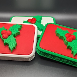 May include: Three rectangular Christmas-themed decorations. Each features a red surface with a green holly and red berry design. The borders are white or green. The decorations are on a dark surface.