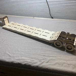 May include: A wooden toy flatbed truck with a long, rectangular bed featuring evenly spaced cut-out slots. The truck has a gooseneck hitch and multiple wheels. The toy is made of light-colored wood.