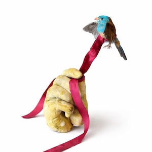 Könnte beinhalten: Eine skurrile Skulptur, die einen Vogel mit blauen und braunen Federn zeigt, der auf einem roten Band sitzt, das um eine strukturierte, gelb getönte Hand gebunden ist. Der Vogel hat teilweise ausgebreitete Flügel, vor weißem Hintergrund.
