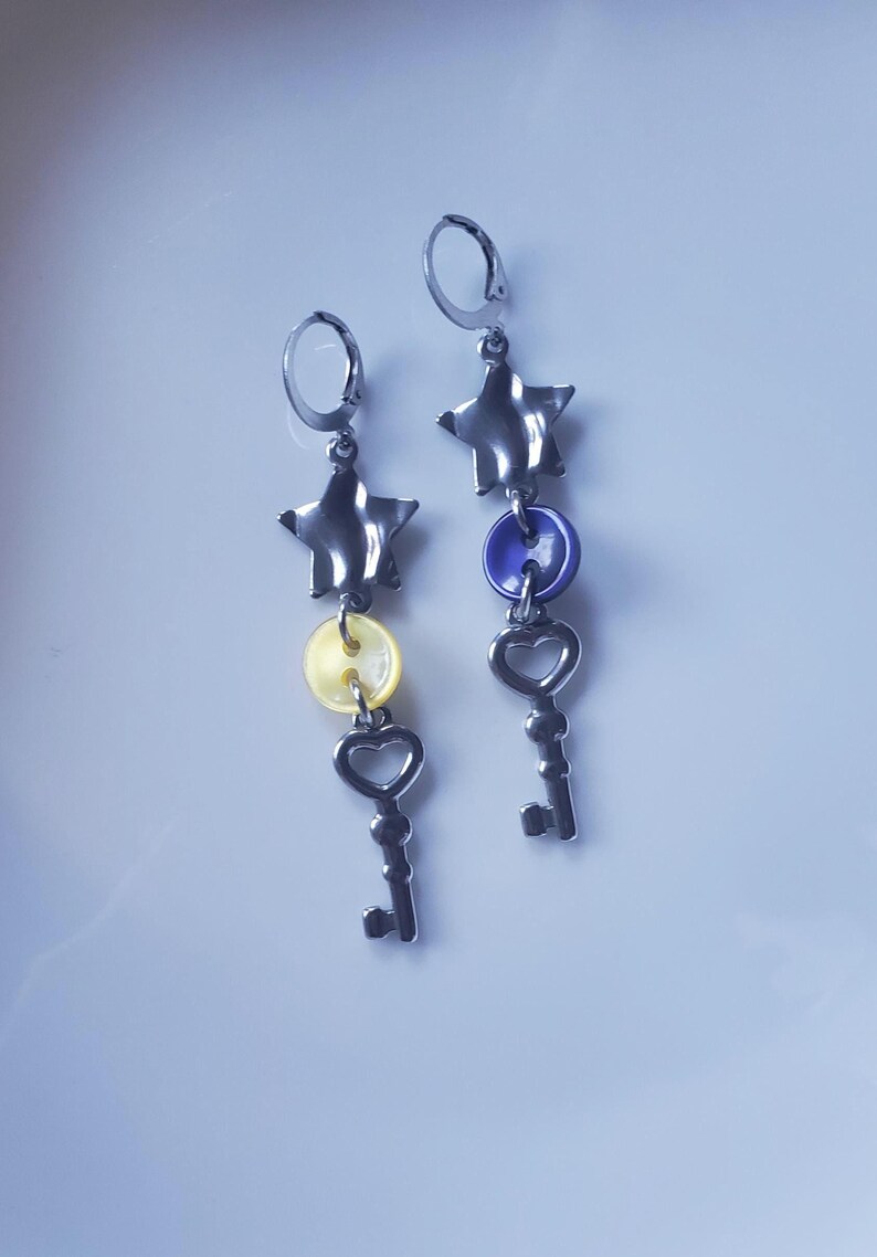 K&ouml;nnte beinhalten: Ein Paar Ohrh&auml;nger mit Stern-, Knopf- und Schl&uuml;sseldesign. Die Ohrringe haben silberfarbene Metallkomponenten, mit einem gelben Knopf an einem Ohrring und einem blauen Knopf am anderen. Die Schl&uuml;ssel haben herzf&ouml;rmige Details.