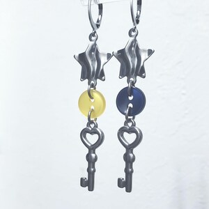 K&ouml;nnte beinhalten: Ein Paar silberfarbene Ohrringe. Jeder Ohrring hat eine sternf&ouml;rmige Oberseite, einen Knopf und einen Schl&uuml;sselanh&auml;nger. Ein Ohrring hat einen gelben Knopf, der andere einen blauen Knopf. Die Schl&uuml;sselanh&auml;nger haben herzf&ouml;rmige Ausschnitte.
