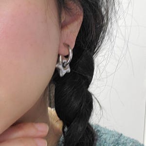 May include: A silver star-shaped earring with a hoop attached to it. The earring is worn in the left ear of a person with long black hair.