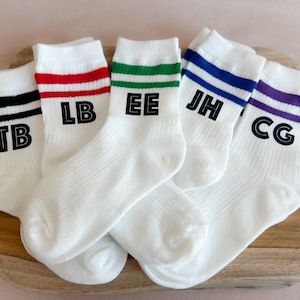 Könnte beinhalten: Fünf Paar weiße Sportsocken mit farbigen Streifen am oberen Rand. Jedes Paar hat einen anderen Farbstreifen: schwarz, rot, grün, blau und lila. Jede Socke hat zwei schwarze Initialen.