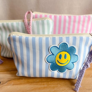 Könnte beinhalten: Drei gestreifte Kosmetiktaschen in Pastellfarben: Blau, Rosa und Grün. Die vordere Tasche hat eine blaue Blume mit einem gelben Smiley-Gesicht. Jede Tasche hat einen Reißverschluss und eine passende geflochtene Handschlaufe.