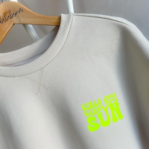 Könnte beinhalten: Ein hellbeiger Pullover mit einem neongrünen Aufdruck, der "Girls Just Wanna Have Sun" lautet.
