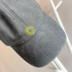 Könnte beinhalten: Eine dunkelgraue Baseballkappe mit einem gestickten gelben Sonnenmotiv.