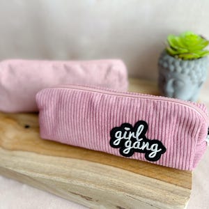 Könnte beinhalten: Zwei rosa Stifteetuis aus Cord. Das vordere Etui hat einen schwarz-weißen Aufnäher mit der Aufschrift "girl gang". Die Etuis liegen auf einem Holzbrett. Eine kleine Sukkulentenpflanze und eine graue Buddha-Kopf-Statue befinden sich im Hintergrund.