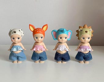 May include: Four small dolls with different animal headbands. Each doll is wearing a pink top, blue jeans, and has a different animal headband: a dog, a deer, an elephant, and a lion.