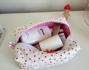 May include: White quilted makeup bag with a red heart pattern. The bag is open and contains various beauty products, including a pink bottle with the word "glossier" on it.