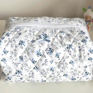 May include: A white quilted makeup bag with a blue floral pattern and a blue ribbon closure. The bag has a white lace trim and a zipper closure.