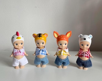 May include: Four small, colorful dolls with different animal headbands. Each doll is wearing a plaid shirt and denim shorts. The dolls are standing on a white surface.