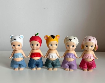 May include: Five small plastic dolls with different animal headbands and outfits. The dolls are wearing blue, purple, pink, and red outfits. The dolls are all smiling.