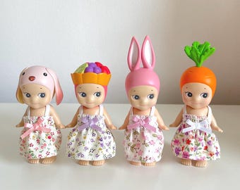 May include: Four small dolls with different headwear and floral print dresses. The dolls are wearing a pink bunny hat, a fruit basket hat, a carrot hat, and a pink dog hat.