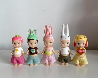 May include: Five small, colorful, plastic dolls with different animal headbands. The dolls are wearing different outfits, including a pink dress, a green dress, a blue dress, a white dress, and a yellow dress. The dolls are all smiling.